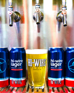 Pic of pint of Hi-Wire Lager draft between four Hi-Wire Lager cans