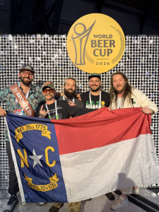 Hi-Wire staff at the 2026 World Beer Cup with smiling faces pose in front of a WBC backdrop while holding the state flag of North Carolina