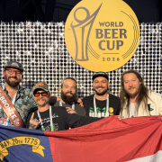Hi-Wire staff at the 2026 World Beer Cup with smiling faces pose in front of a WBC backdrop while holding the state flag of North Carolina
